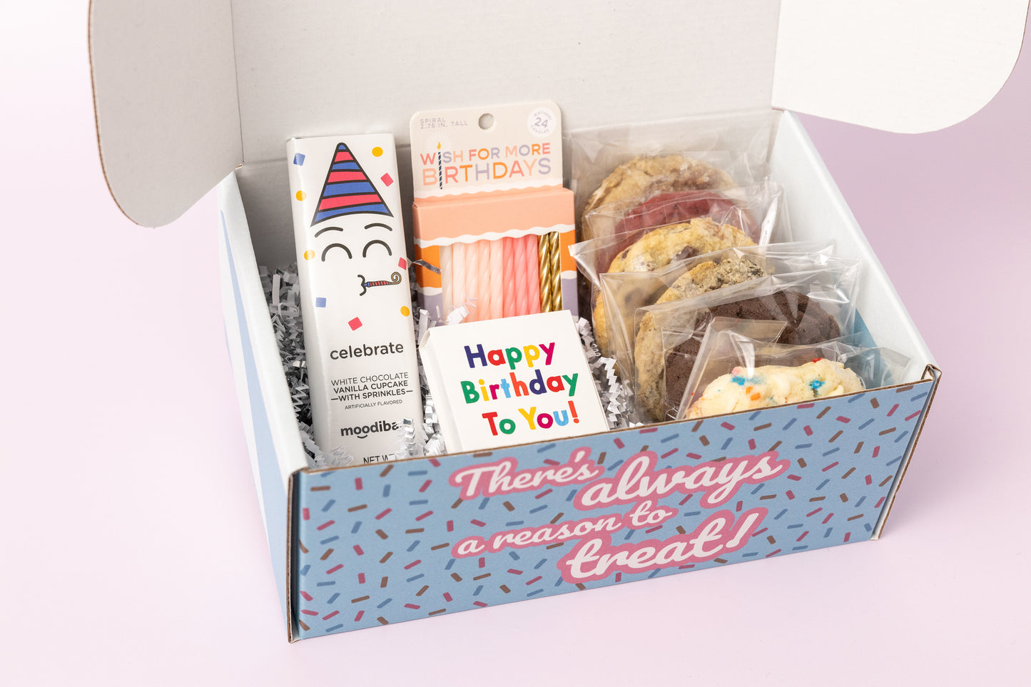 Birthday-themed gift box featuring individually wrapped cookies, a "Celebrate" white chocolate vanilla cupcake bar, pink spiral birthday candles, and a "Happy Birthday to You" matchbox. The box reads, "There's always a reason to treat!" with shredded paper filler and a light pink background.