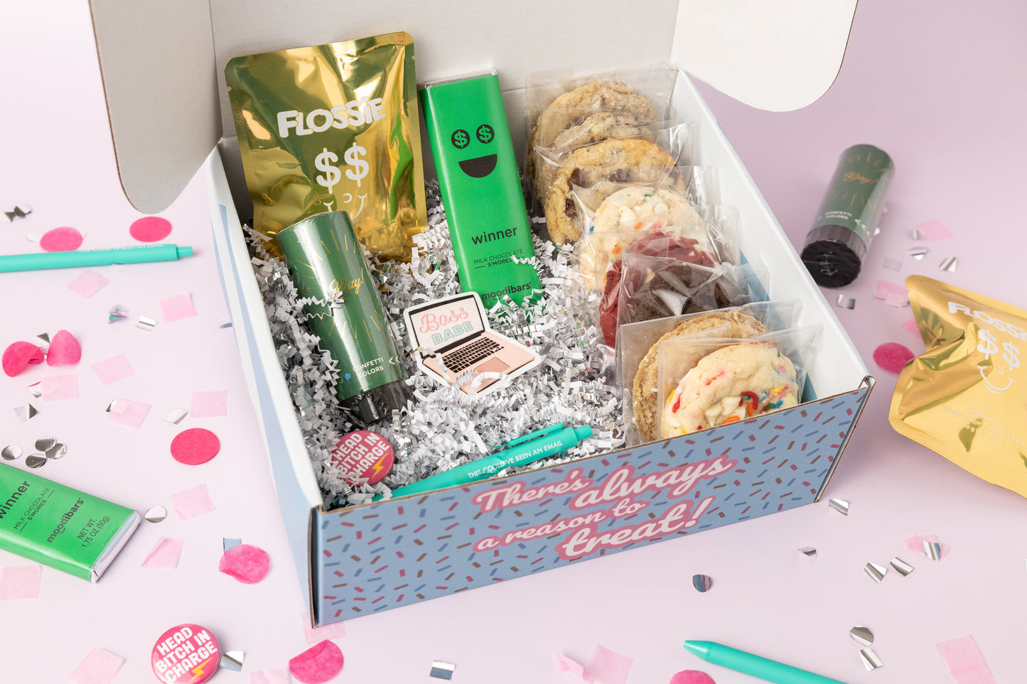 Boss-themed gift box featuring individually wrapped cookies, a "Winner" milk chocolate mint bar, Flossie gold candy pouch, a green confetti popper, a "Boss Babe" laptop sticker, and pins with phrases like "Head Bitch in Charge." The box reads, "There's always a reason to treat!" with pink confetti and a teal pen decorating the background.