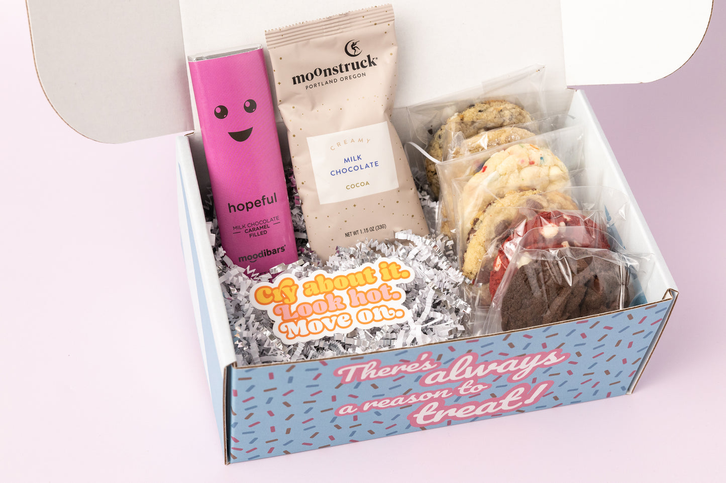 Breakup-themed gift box featuring individually wrapped cookies, a "Hopeful" milk chocolate caramel bar, a packet of creamy milk chocolate cocoa, and a sticker with the phrase "Cry About It. Look Hot. Move On." The box reads, "There's always a reason to treat!" with shredded paper filler and a light pink background.