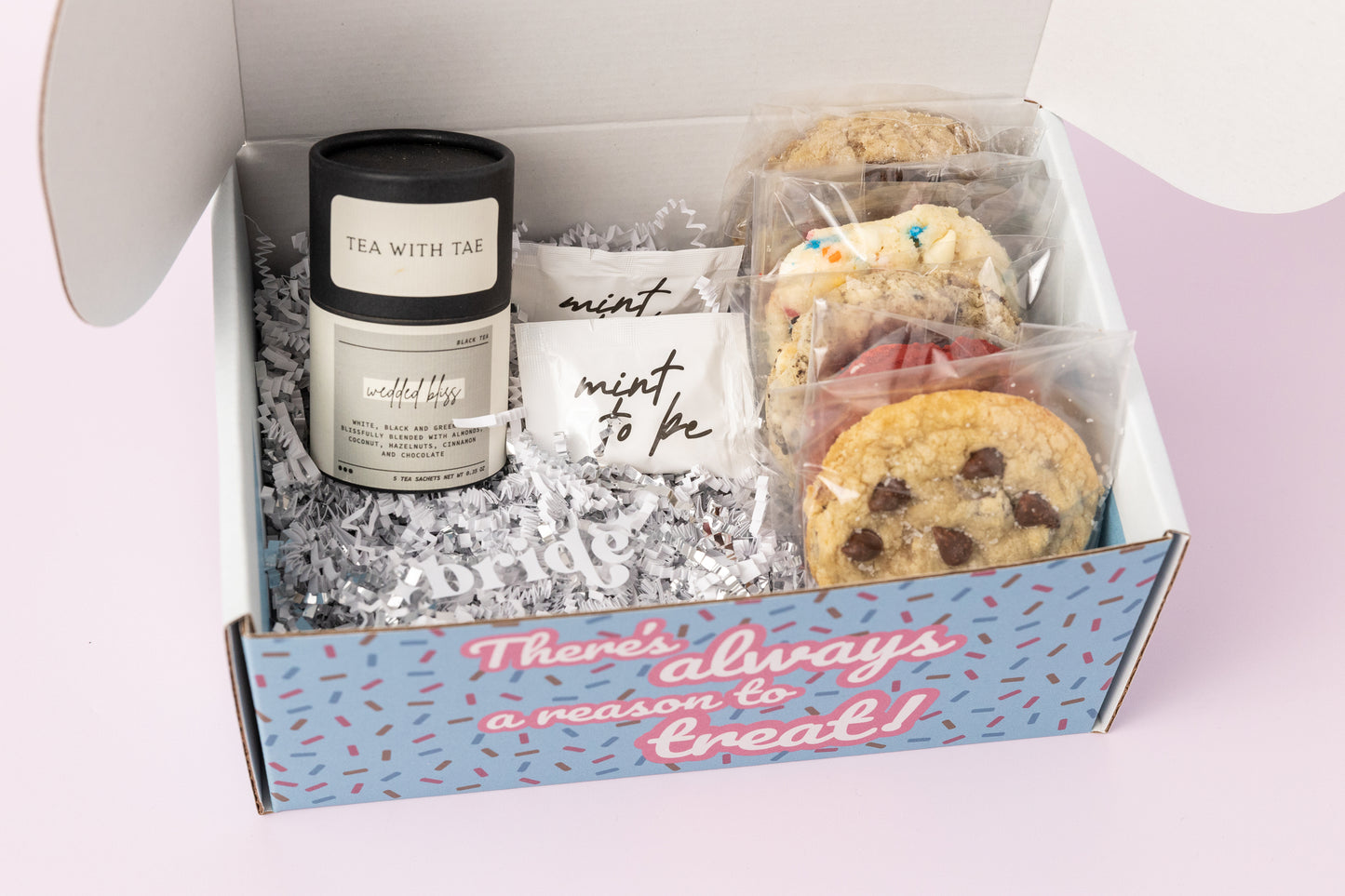 Bridal-themed gift box featuring individually wrapped cookies, a container of "Wedded Bliss" black tea from Tea with Tae, and "Mint to Be" mint packets. The box reads, "There's always a reason to treat!" with shredded paper filler and a light pink background.