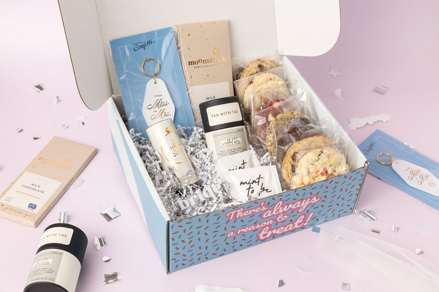 Bridal-themed gift box featuring individually wrapped cookies, a "From Miss to Mrs." keychain, a milk chocolate bar, Tea with Tae packets, a confetti popper, and "Mint to Be" mints. The box reads, "There's always a reason to treat!" with shredded paper filler and confetti decorations on a light pink background.