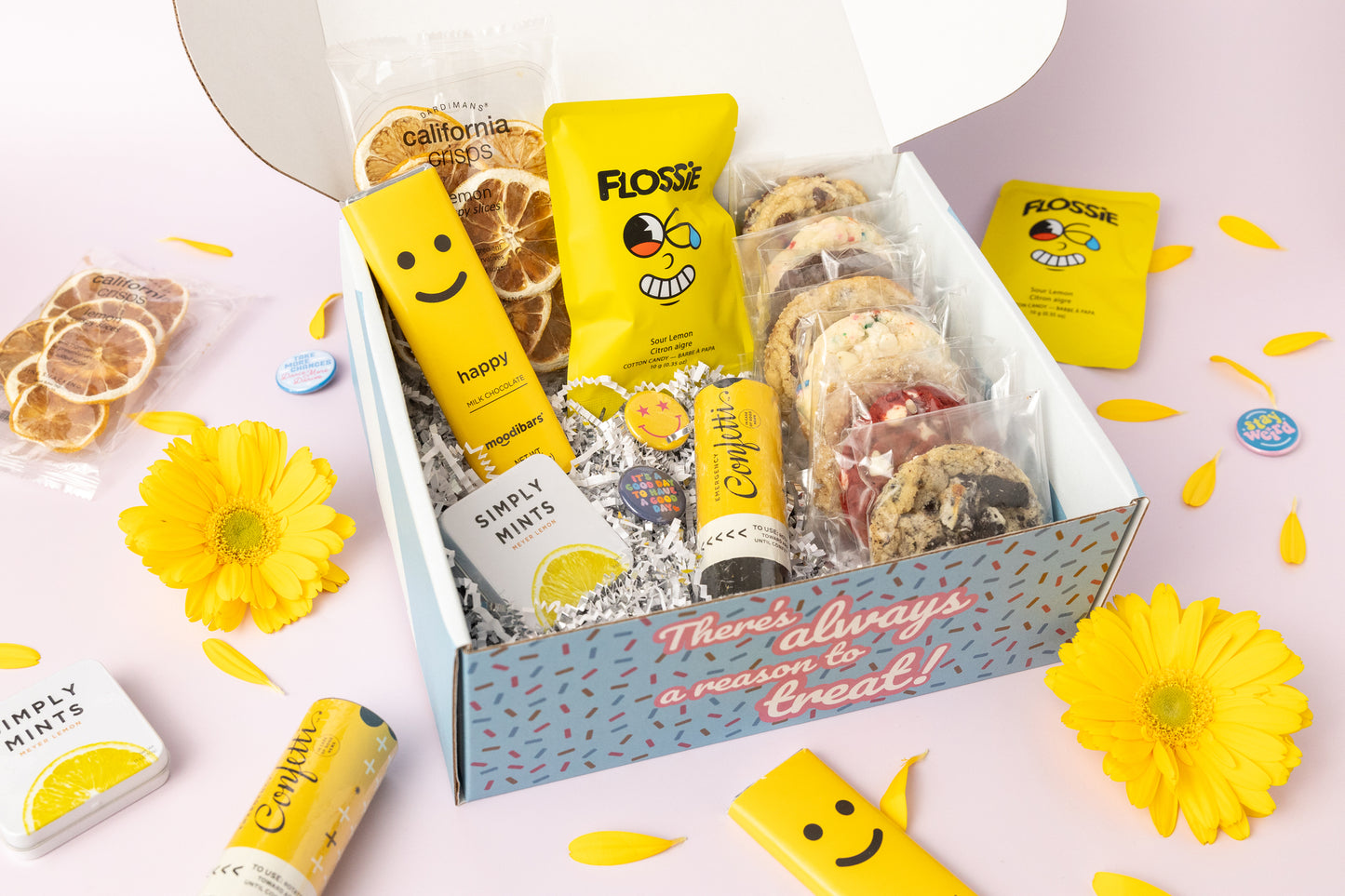 Lemon-themed gift box featuring individually wrapped cookies, dried lemon slices, a yellow "Happy" chocolate bar, Flossie sour lemon candy, Simply Mints, and a confetti popper. The box reads, "There's always a reason to treat!" with yellow flower petals and daisies decorating the pink background.