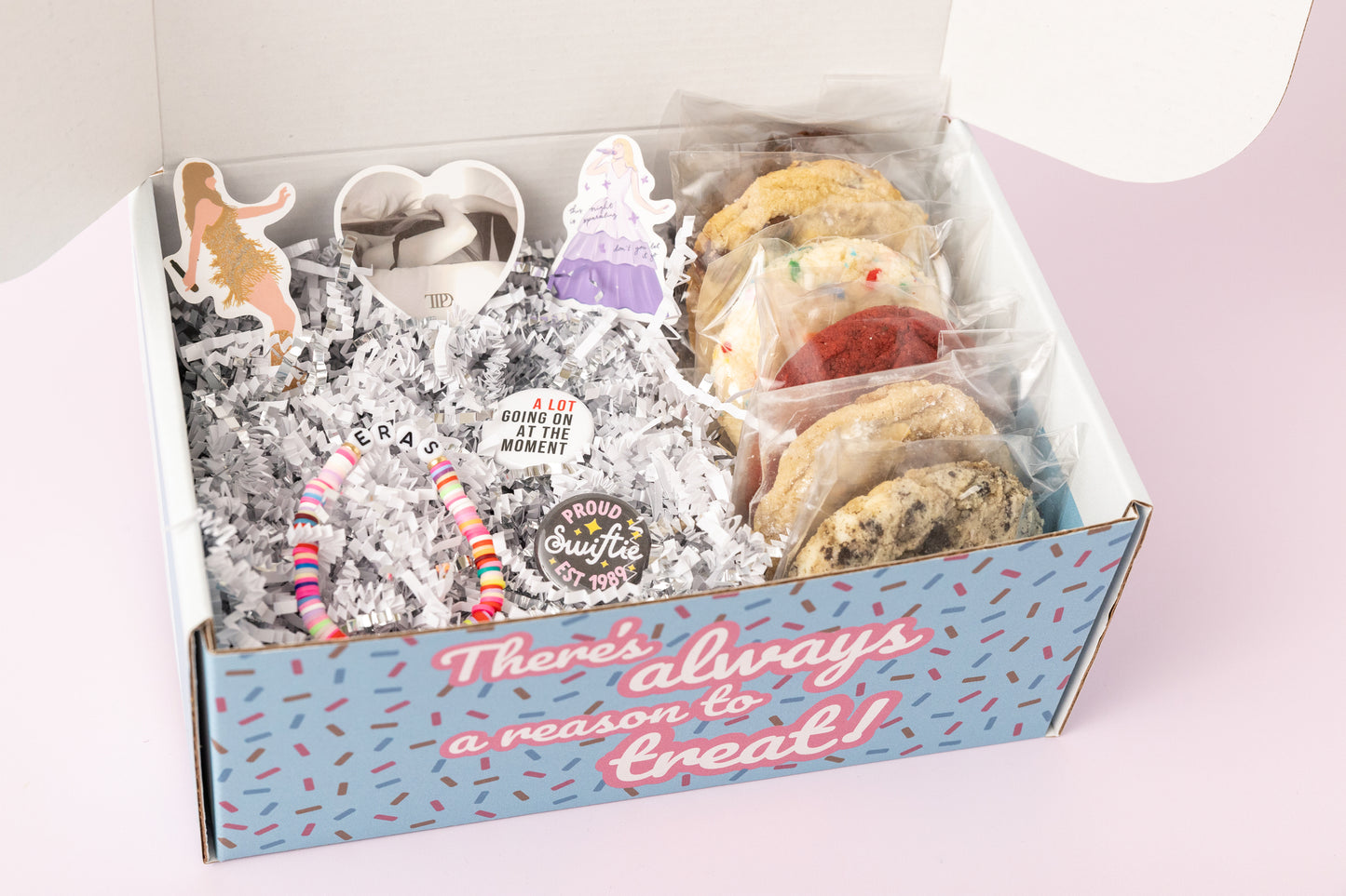 Taylor Swift-themed gift box featuring individually wrapped cookies, colorful "Eras" beaded bracelets, Swiftie pins, and stickers, including a heart-shaped design and images of Taylor. The box says, "There's always a reason to treat!" with shredded paper filler and a pink background.