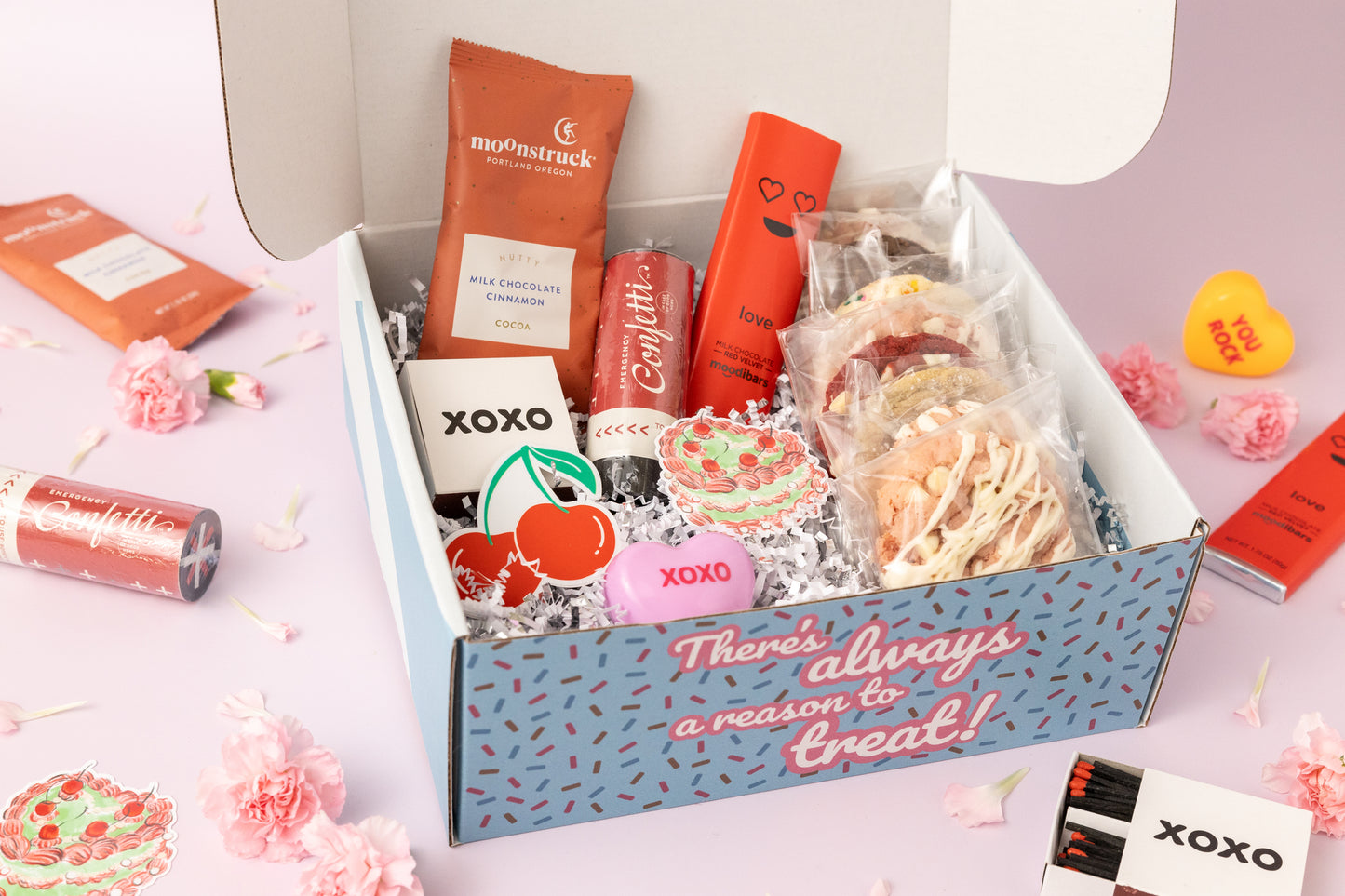 Open Valentine's Day gift box featuring assorted cookies in individual packaging, gourmet hot cocoa mix, a red chocolate bar with a heart design, stickers with cherry and "XOXO" motifs, and festive extras like a confetti popper. The box is surrounded by pink carnations and a light pink background with additional Valentine's Day-themed decor. The box exterior reads, "There's always a reason to treat!"