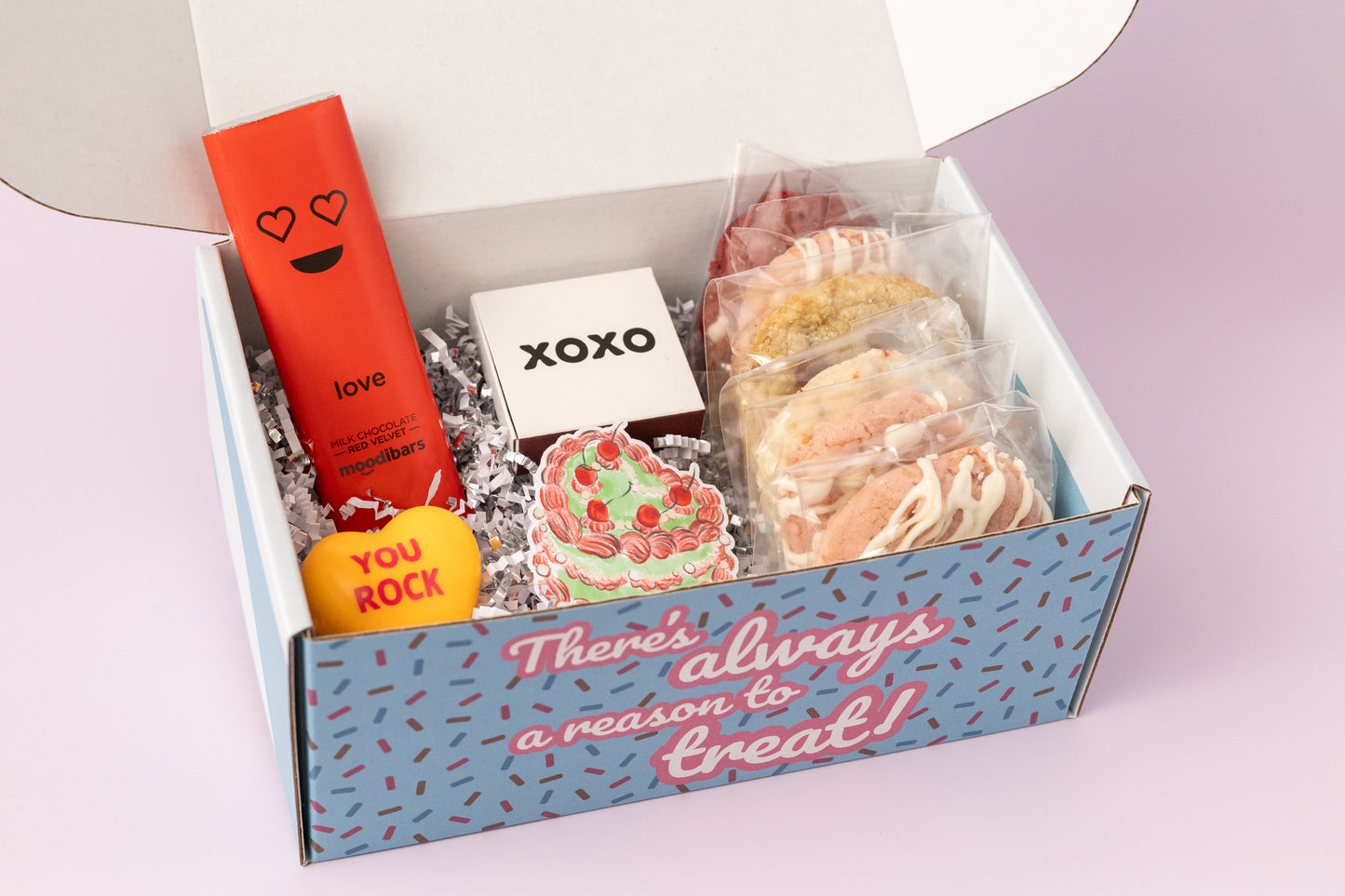 Valentine's Day gift box featuring individually packaged cookies in various flavors, a red chocolate bar labeled "Love," a small white box with "XOXO," a yellow heart-shaped item reading "YOU ROCK," and a decorative sticker with a heart-shaped cookie design. The box exterior displays the phrase "There's always a reason to treat!" against a blue background with confetti sprinkles. The contents rest on festive shredded paper, with a soft pink background enhancing the romantic theme.