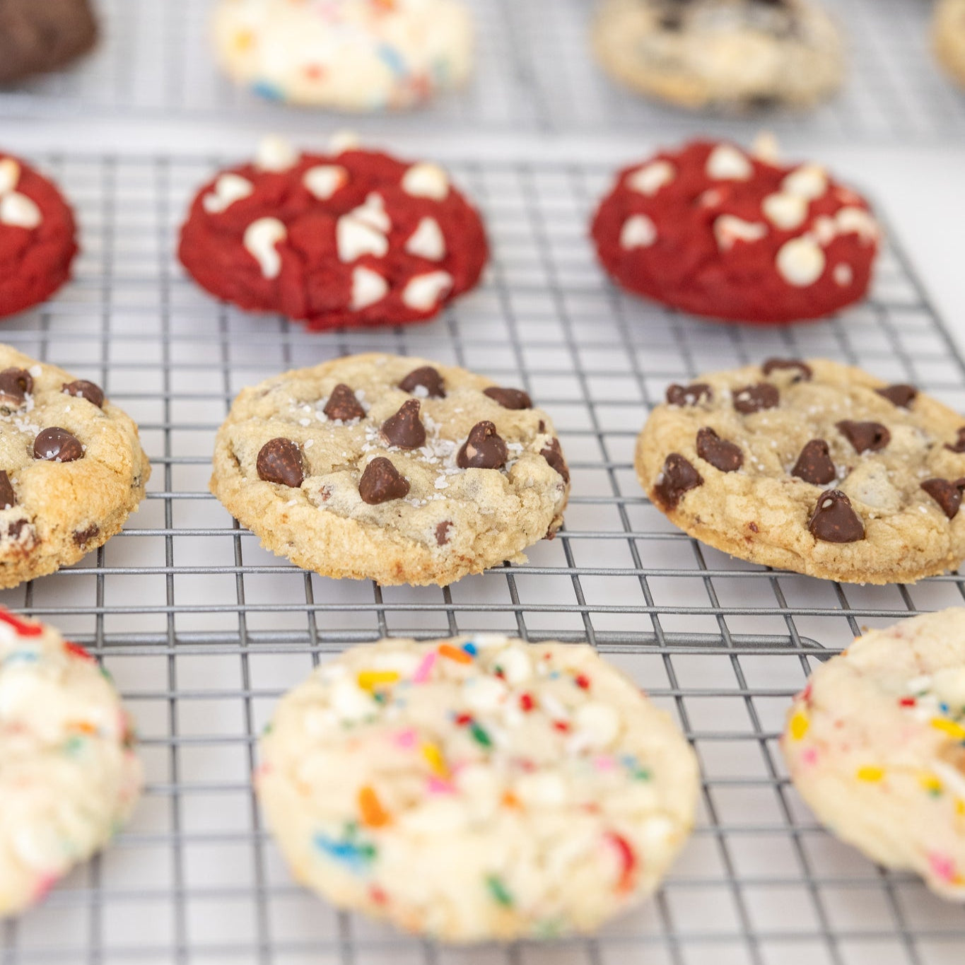 Gourmet cookies cooling on a wire rack. Flavors shown are red velvet, chocolate chip, and confetti cake.