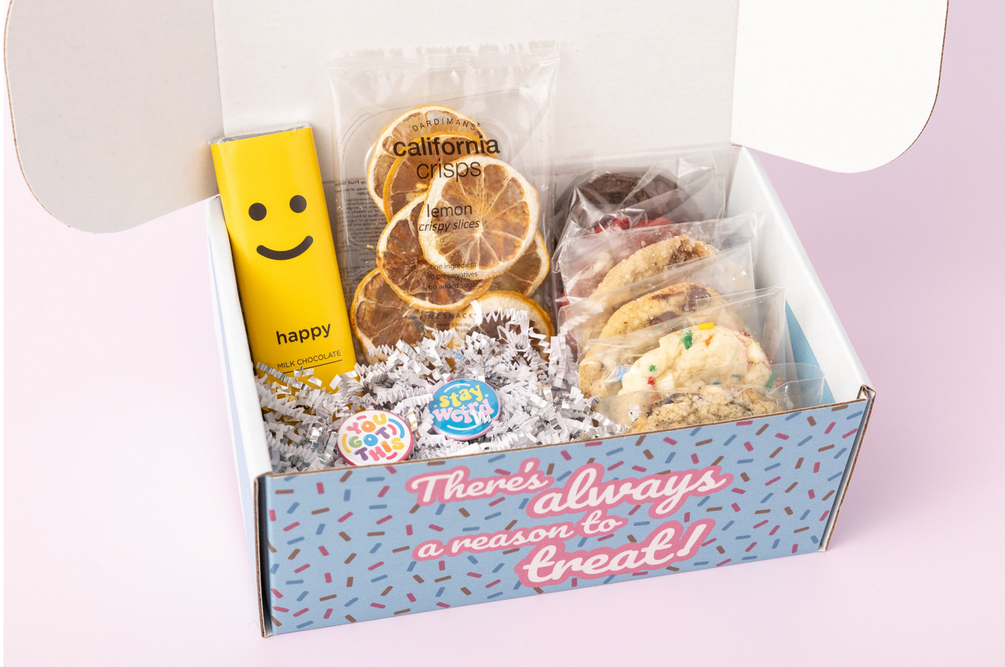 Lemon-themed gift box featuring individually wrapped cookies, a yellow "Happy" chocolate bar, dried lemon slices, and motivational pins with phrases like "You Got This" and "Stay Weird." The box reads, "There's always a reason to treat!" with shredded paper filler and a light pink background.