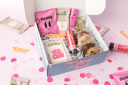 Breakup-themed gift box featuring individually wrapped cookies, Flossie pink vanilla candy, a milk chocolate cocoa packet, a confetti popper, and motivational stickers with phrases like "You're Doing Amazing Sweetie" and "Cry About It, Look Hot, Move On." The box reads, "There's always a reason to treat!" with pink confetti scattered on a light pink background.