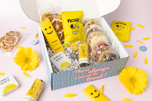 Lemon-themed gift box featuring individually wrapped cookies, dried lemon slices, a yellow "Happy" chocolate bar, Flossie sour lemon candy, Simply Mints, and a confetti popper. The box reads, "There's always a reason to treat!" with yellow flower petals and daisies decorating the pink background.