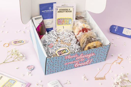 Mama-themed gift box featuring individually wrapped cookies, a chocolate bar, a packet of dark chocolate cocoa, "MAMA" and "MOM" stickers, a "MAMA" keychain, and a gold "MAMA" necklace. The box reads, "There's always a reason to treat!" with shredded paper filler and baby’s breath flowers decorating the pink background.