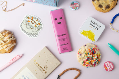 Assorted snacks and products including a pink 'hopeful' bar, colorful cookies, and a 'Simply Mints' tin on a light surface.