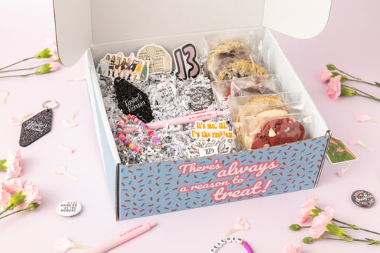 Taylor Swift-themed gift box featuring individually wrapped cookies, a "Taylor's Version" keychain, beaded bracelets, a pink pen reading "In My Swiftie Era," and Swiftie pins and stickers. The box says, "There's always a reason to treat!" and includes shredded paper filler with pink carnations decorating the background.
