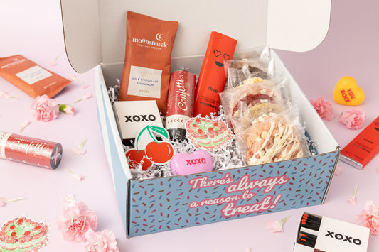 Open Valentine's Day gift box featuring assorted cookies in individual packaging, gourmet hot cocoa mix, a red chocolate bar with a heart design, stickers with cherry and "XOXO" motifs, and festive extras like a confetti popper. The box is surrounded by pink carnations and a light pink background with additional Valentine's Day-themed decor. The box exterior reads, "There's always a reason to treat!"
