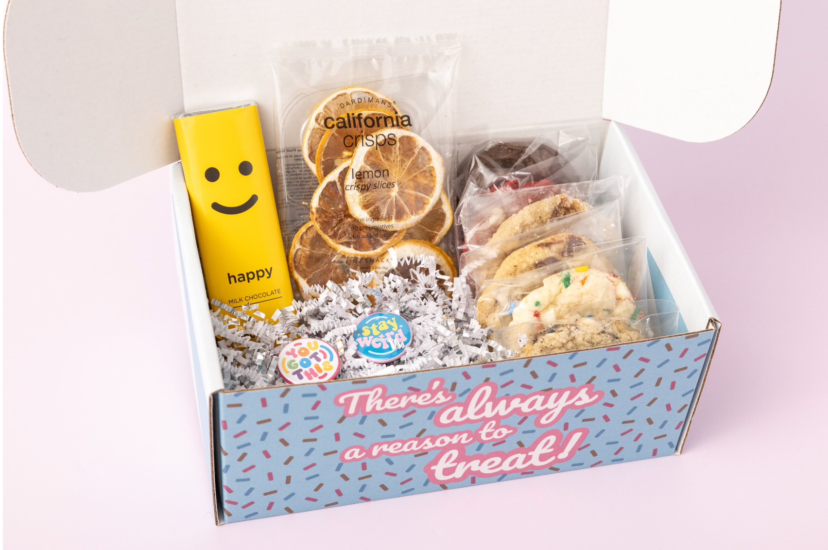 Lemon-themed treat box featuring individually wrapped cookies, a yellow "Happy" chocolate bar, dried lemon slices, and motivational pins with phrases like "You Got This" and "Stay Weird." The box reads, "There's always a reason to treat!" with shredded paper filler and a light pink background.